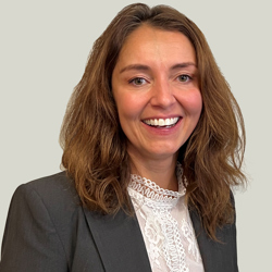 A person with shoulder-length wavy brown hair is wearing a dark gray blazer over a white lace blouse. The background is plain and light-colored.