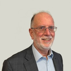 A person wearing glasses and with a light coloured beard, wearing a grey checkered suit jacket over a light blue striped dress shirt, set against a plain light-colored background