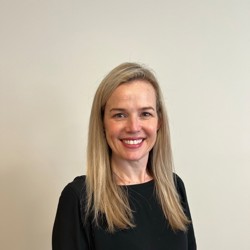 A person with long, straight blonde hair standing against a plain beige background, wearing a black long-sleeved top and dark pants.