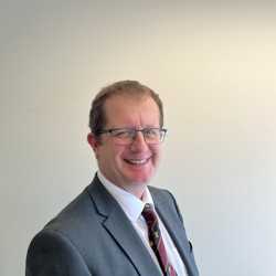 A person wearing a grey suit jacket, white dress shirt, and a colourful striped tie with small yellow and blue designs. The background is a plain light-coloured wall.