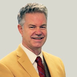 A person wearing a mustard yellow blazer, white dress shirt, red and blue striped tie, and a dark grey vest. The background is plain light grey.