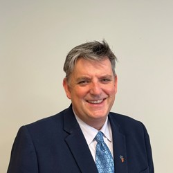 A person wearing a dark blue suit, white dress shirt, and a light blue patterned tie. The background is plain and light-coloured.