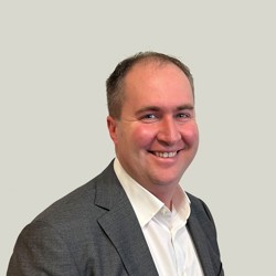 A person wearing a white dress shirt and a dark gray suit jacket, seated against a plain light gray background. 