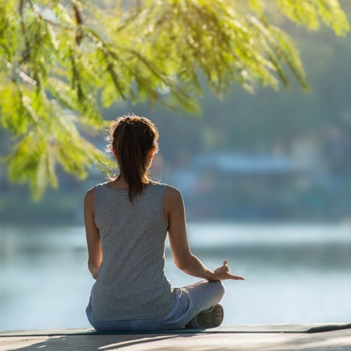Woman sitting with legs crossed looking out to a lake while sitting under a tree.