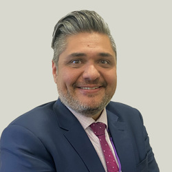 A person wearing a dark blue suit, light pink checkered dress shirt, and a red patterned tie. The person has short, neatly styled hair and is standing against a plain light grey background.