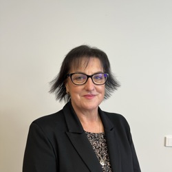 A person with short, dark hair standing against a plain light-coloured wall. The person is wearing glasses, a black blazer, and a patterned blouse.