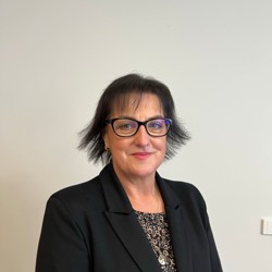 A person with short, dark hair standing against a plain light-coloured wall. The person is wearing glasses, a black blazer, and a patterned blouse.