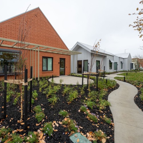 Modern healthcare or community facility with red brick and light grey buildings, landscaped garden beds, young trees, and a curved accessible pathway leading to the entrance.