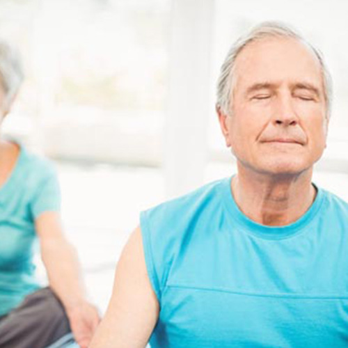 Gentleman sitting in the foreground in a blue top, with lady sitting in the background in a blue top. Both are crossed legged and sitting with eyes closed meditating.