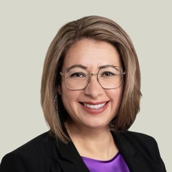 A person with shoulder-length light brown hair wearing glasses, a black blazer, and a purple blouse. The background is plain and light-coloured.