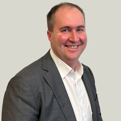 A person wearing a white dress shirt and a dark gray suit jacket, seated against a plain light gray background. 