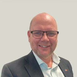 A person wearing a dark suit jacket over a light blue dress shirt. The background is plain and light grey. A small pin is visible on the left lapel of the suit jacket.