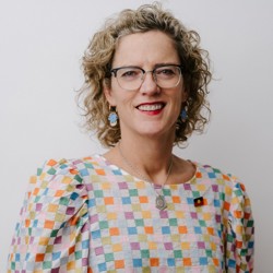 Portrait of a person with curly hair and glasses wearing a colourful checkered blouse, standing against a light grey background.