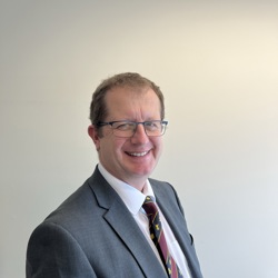 A person wearing a grey suit jacket, white dress shirt, and a colourful striped tie with small yellow and blue designs. The background is a plain light-coloured wall.
