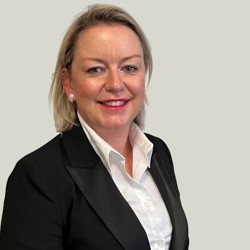 A person with shoulder-length blonde hair wearing a black blazer over a white dress shirt. The background is plain light gray.