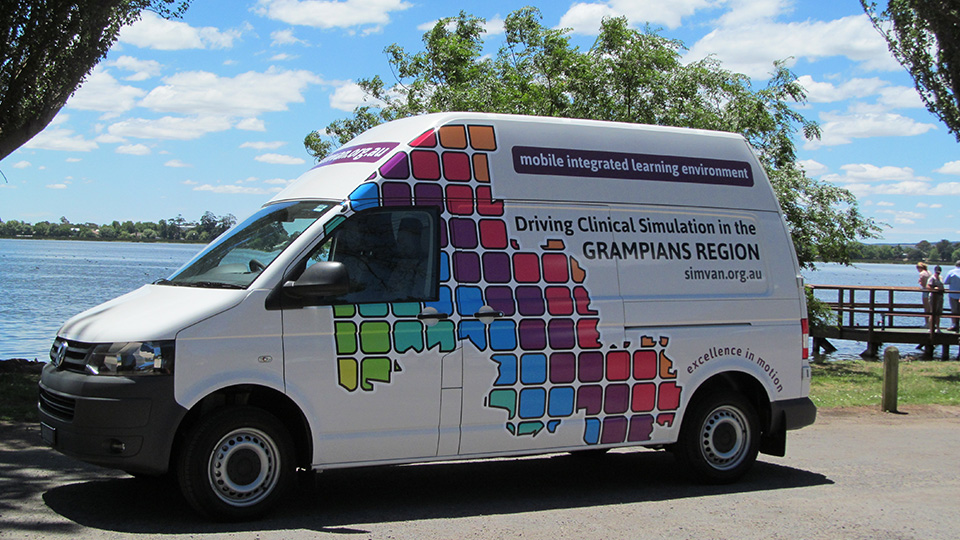 Simvan White van parked near a lake with colorful square graphics on the side forming a map-like design. Text on the van reads: 'Mobile integrated learning environment. Driving Clinical Simulation in the Grampians Region. simvan.org.au. Excellence in motion.' Trees and a wooden pier are visible in the background under a bright blue sky with scattered clouds.