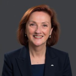 Professional headshot of a person wearing a dark navy blazer and matching top, facing the camera against a dark grey background.