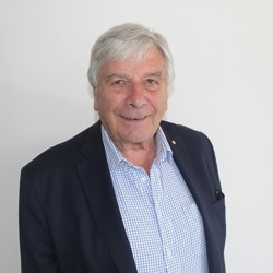 A person with grey hair is wearing a dark blazer over a light blue checkered shirt. The background is plain and white.