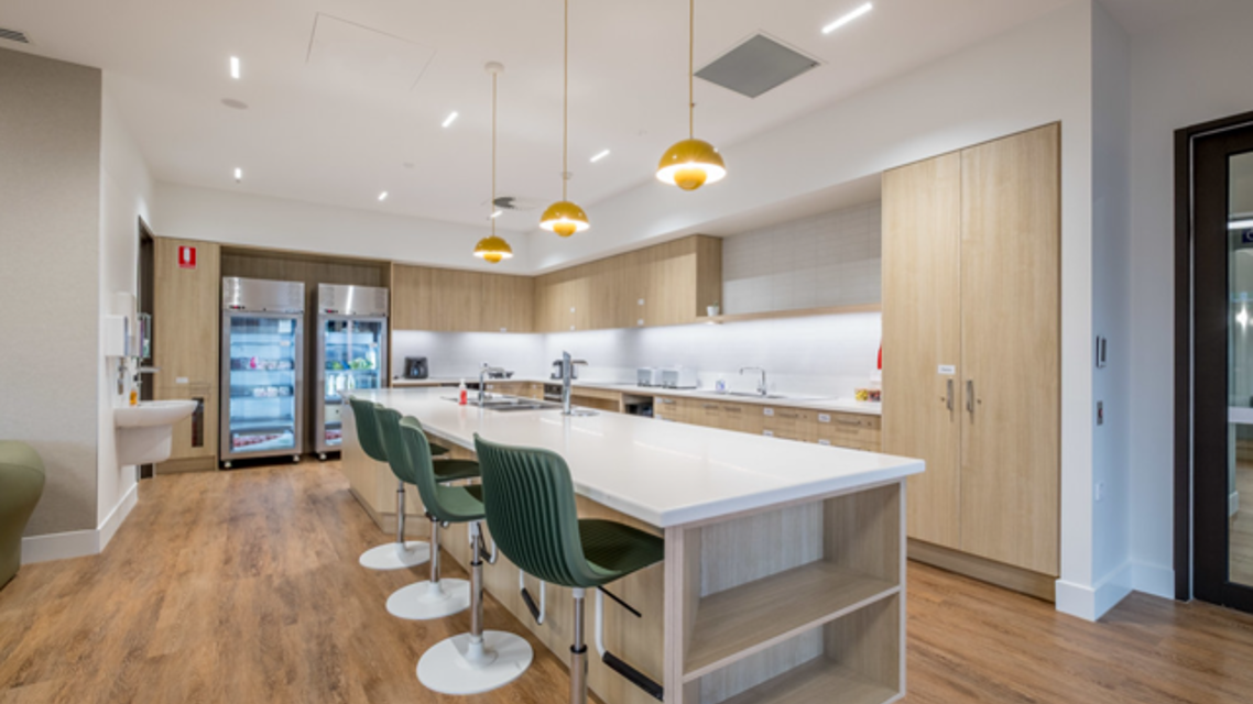 Modern communal kitchen with a large white island bench, four green bar stools, light timber cabinetry, timber-look flooring, and three pendant lights. Refrigerated beverage units and stainless-steel appliances are positioned along the back wall in a bright, contemporary space.