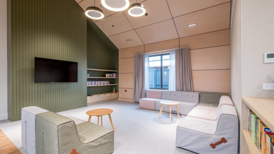 Contemporary lounge and relaxation room featuring soft modular seating, a wall-mounted television, built-in shelving, and small round tables. The space includes a green feature wall, timber wall panels, large window with curtains, and circular pendant lights, creating a calm and welcoming environment.