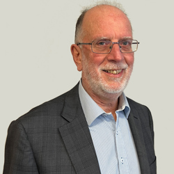 A person wearing glasses and with a light coloured beard, wearing a grey checkered suit jacket over a light blue striped dress shirt, set against a plain light-colored background