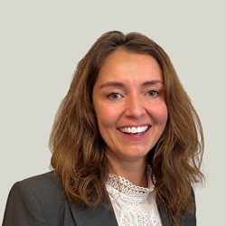 A person with shoulder-length wavy brown hair is wearing a dark gray blazer over a white lace blouse. The background is plain and light-colored.