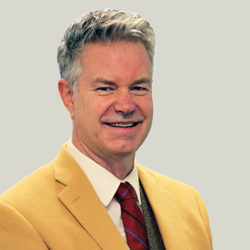 A person wearing a mustard yellow blazer, white dress shirt, red and blue striped tie, and a dark grey vest. The background is plain light grey.