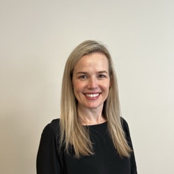 A person with long, straight blonde hair standing against a plain beige background, wearing a black long-sleeved top and dark pants.