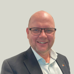 A person wearing a dark suit jacket over a light blue dress shirt. The background is plain and light grey. A small pin is visible on the left lapel of the suit jacket.