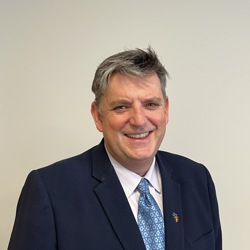 A person wearing a dark blue suit, white dress shirt, and a light blue patterned tie. The background is plain and light-coloured.