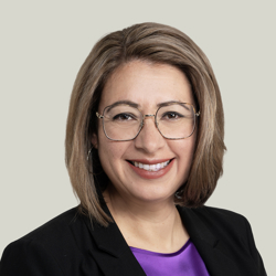 A person with shoulder-length light brown hair wearing glasses, a black blazer, and a purple blouse. The background is plain and light-coloured.