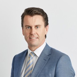 Professional headshot of a person wearing a light blue suit jacket, white shirt, and striped tie against a light grey background.