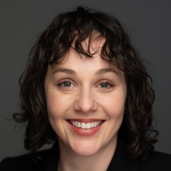 Person wearing a black blazer over a light-colored top, with shoulder-length curly hair, posed against a plain dark background.