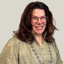 A person with shoulder-length dark hair wearing a light green blouse with puffed sleeves and floral embroidery along the button placket. The background is plain and light-colored.